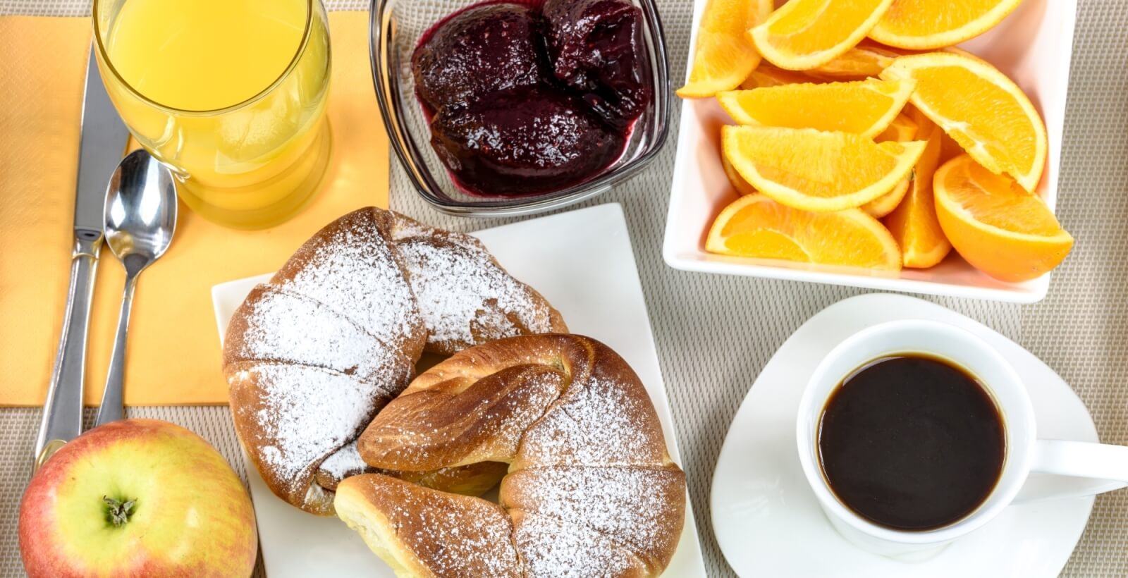 Impostazione della colazione con succo d'arancia, croissant, frutta e caffè su un sottopiatto beige.