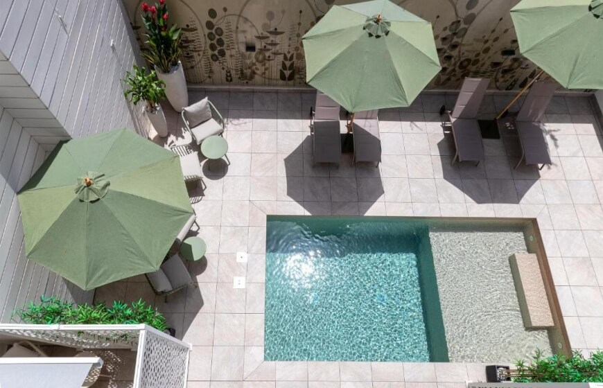 Vista aerea di una piscina rettangolare turchese circondata da un patio con ombrelloni verdi e piante in vaso.