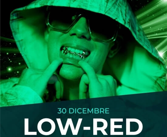 Persona con giacca e occhiali da sole, dita sulle bretelle, sfondo scuro con luci verdi e testo "30 DICEMBRE LOW-RED".