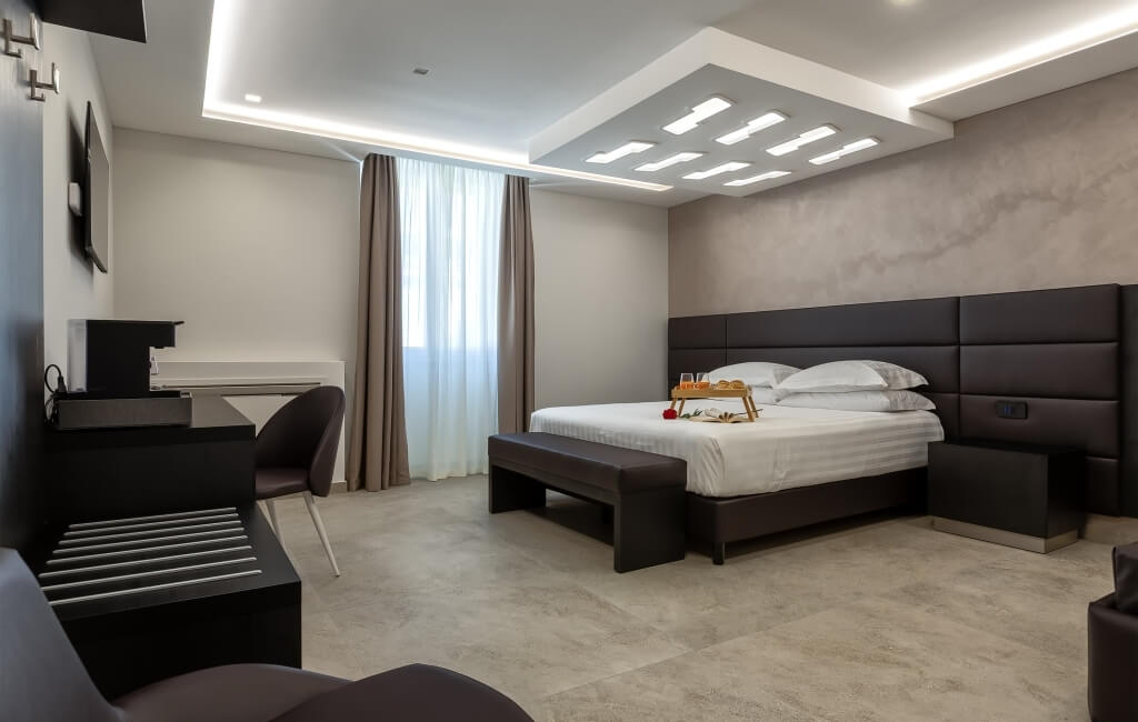 Camera d'hotel moderna con letto grande, scrivania, TV e pavimento in piastrelle chiare. Pareti beige e accento marmoreo.