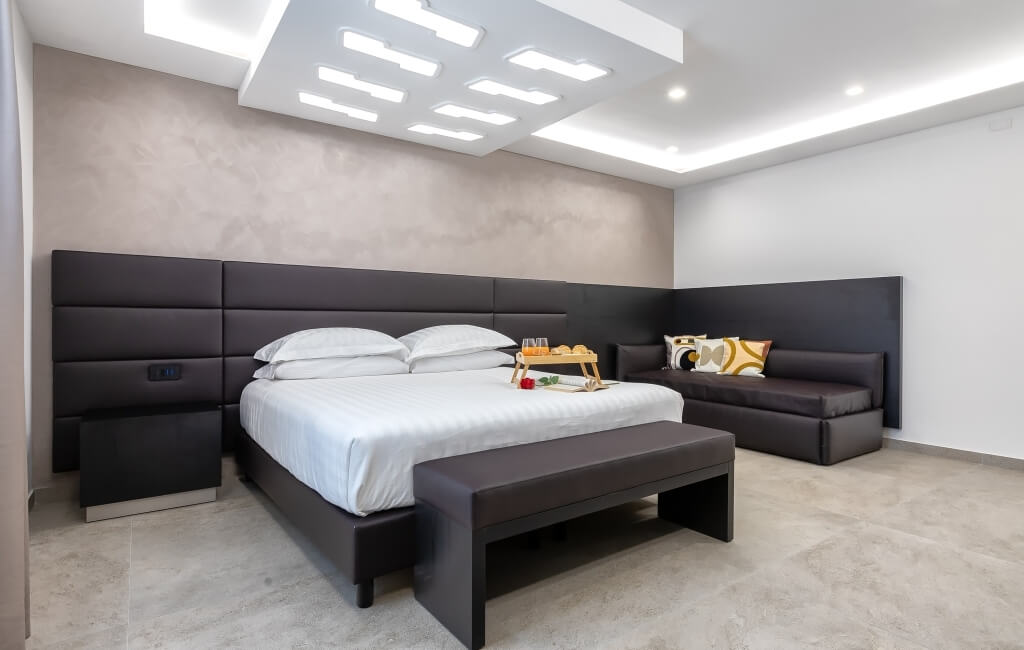 Camera minimalista con letto grande, testiera in pelle marrone, cuscini bianchi e panchina in pelle marrone ai piedi.