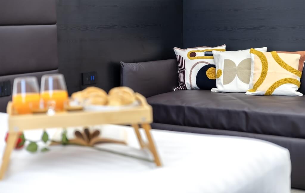 Salotto moderno e accogliente con divano grigio, cuscini decorativi e tavolo imbandito per colazione con succhi e dolci.