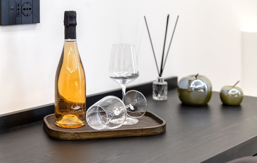 Piano in legno scuro con vino, bicchiere, diffusore e oggetti decorativi su sfondo bianco.