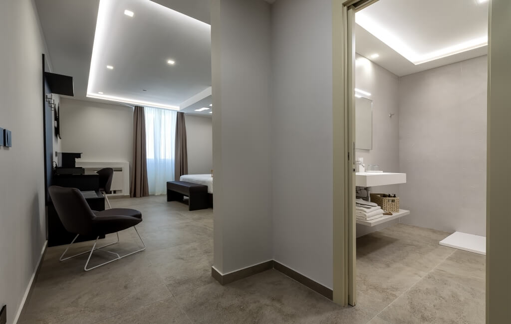 Camera d'hotel moderna e luminosa con bagno, letto, scrivania, TV e sedie nere. Atmosfera pulita e minimalista.