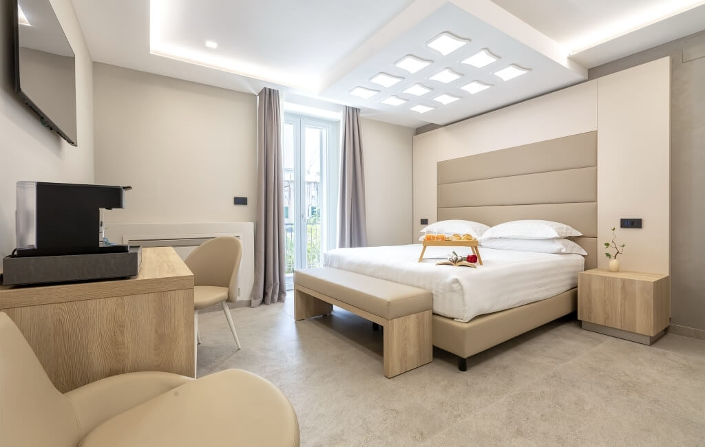 Camera d'hotel moderna con letto beige, biancheria bianca, panca, scrivania e luce naturale.