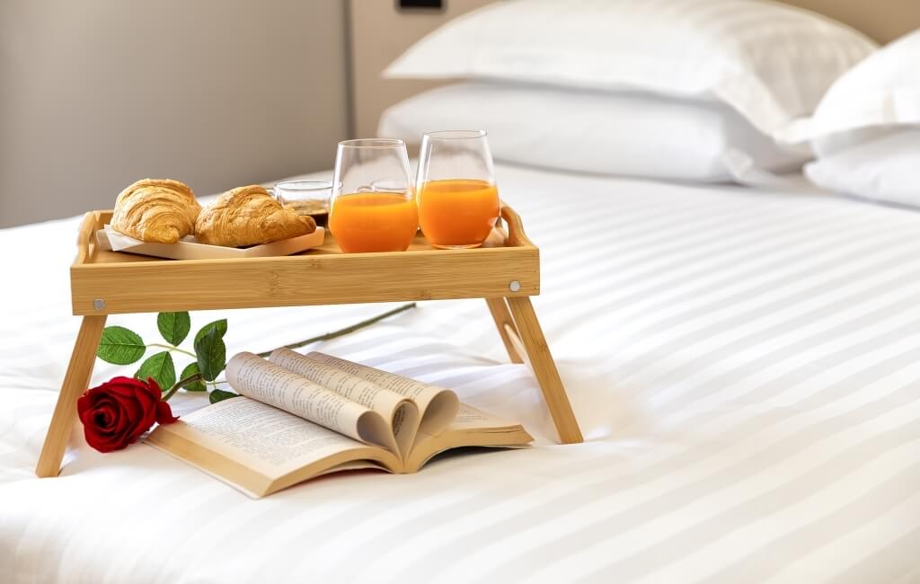 Colazione serena a letto: vassoio di legno con croissant, succo d'arancia, libro aperto e rosa rossa.