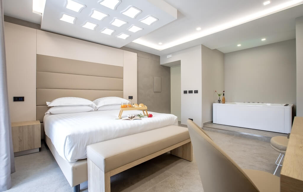 Camera d'hotel moderna con letto grande, vassoio con succhi e dolci, bagno con vasca e champagne. Muri beige e soffitto illuminato.