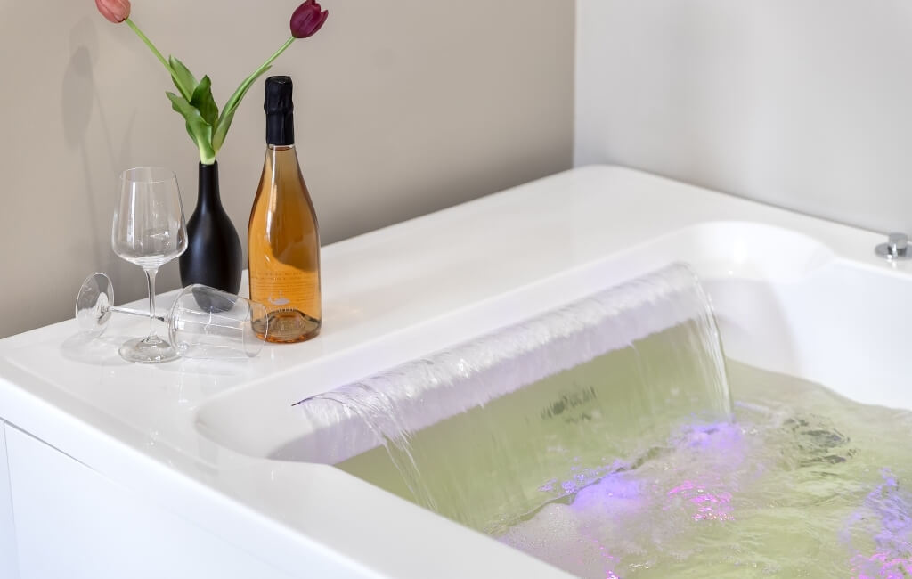 Vasca bianca con acqua verde-gialla, vino e bicchieri, tulipani rosa e viola, sfondo bianco.