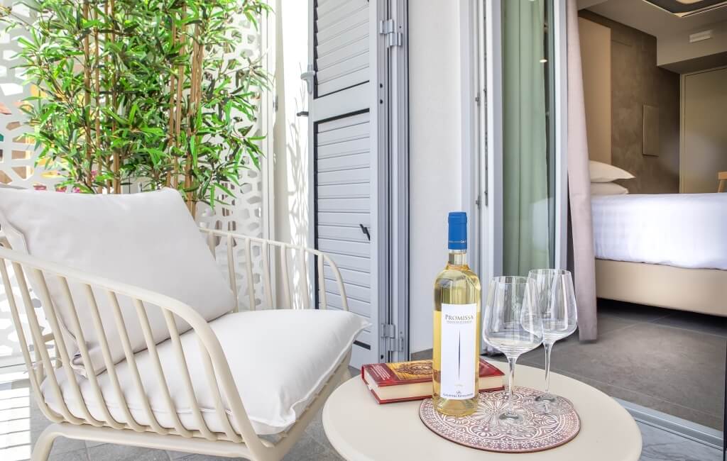 Balcone sereno con sedia bianca, tavolo rotondo, vino, bicchieri e libro rosso, davanti a una porta scorrevole aperta.