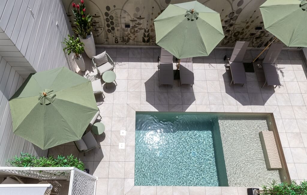 Piscina blu con sdraio e ombrelloni in uno spazio esterno, decorato con un muro bianco e un murale colorato.