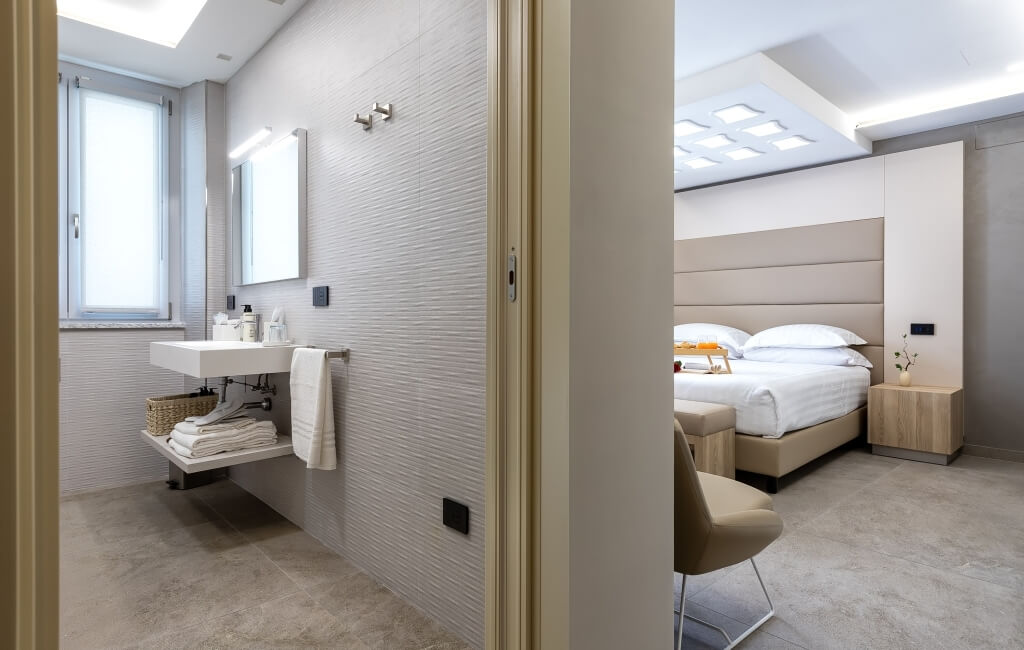 Camera d'hotel moderna con bagno a sinistra e camera da letto a destra, dotata di finestra e arredi eleganti.