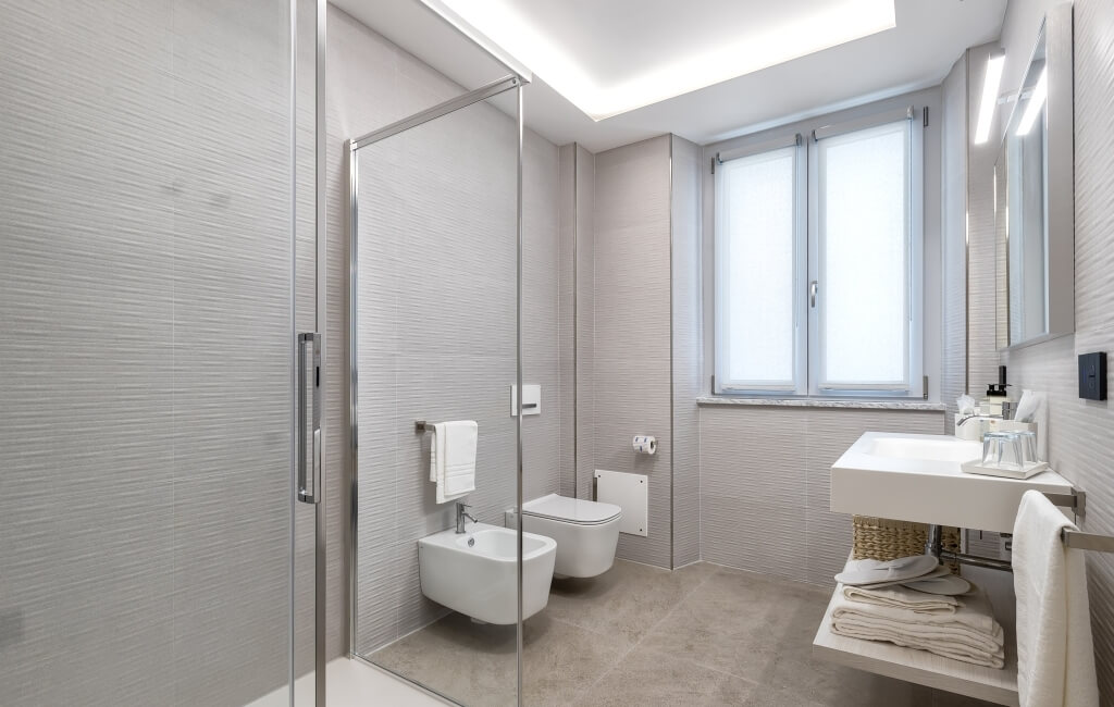 Bagno moderno minimalista con doccia in vetro, wc in angolo, finestra e lavabo bianco. Illuminato da un lucernario.