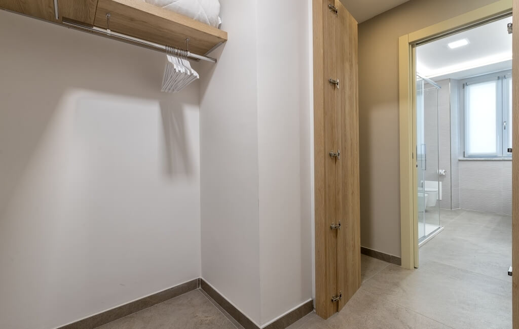 Camera d'hotel con armadio a muro e bagno, pareti bianche e pavimento in legno chiaro, luce naturale nella stanza.