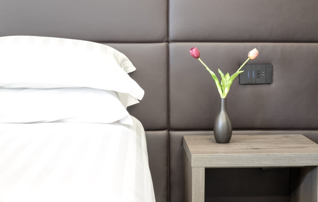 Camera serena con letto bianco, comodino in legno chiaro e vaso grigio con tulipani, outlet dietro il comodino.