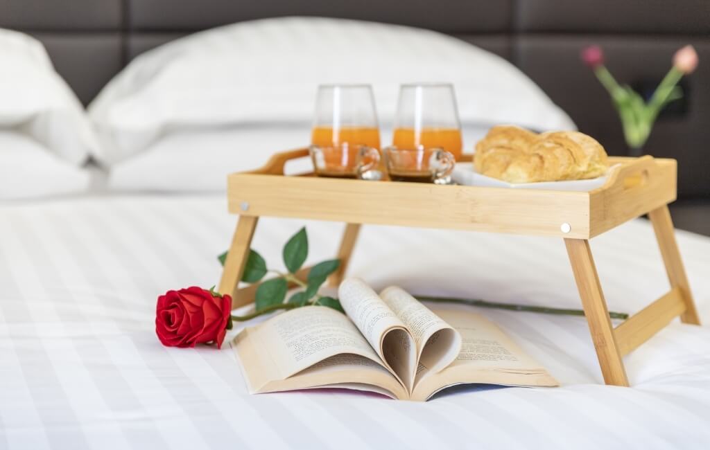 Vassoio da colazione in legno su un letto, con succo d'arancia, croissant, libro aperto e una rosa rossa.