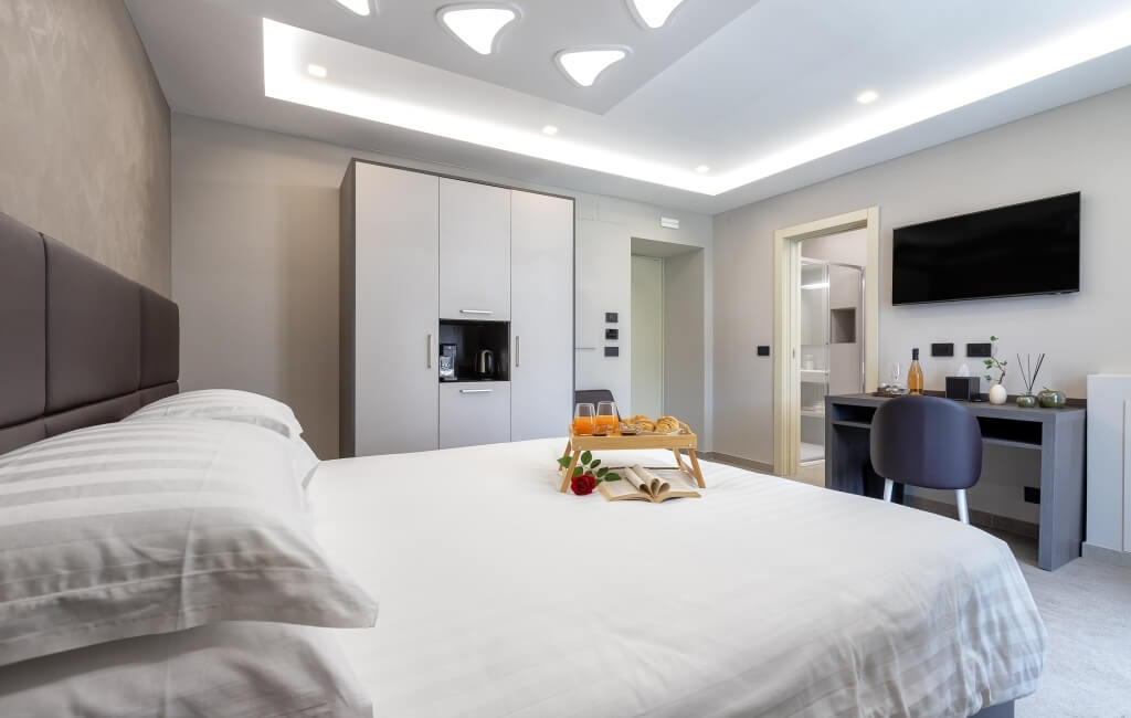 Camera d'hotel moderna con letto, scrivania, TV, e un vassoio con libro, rosa, succo d'arancia e pasticcini.