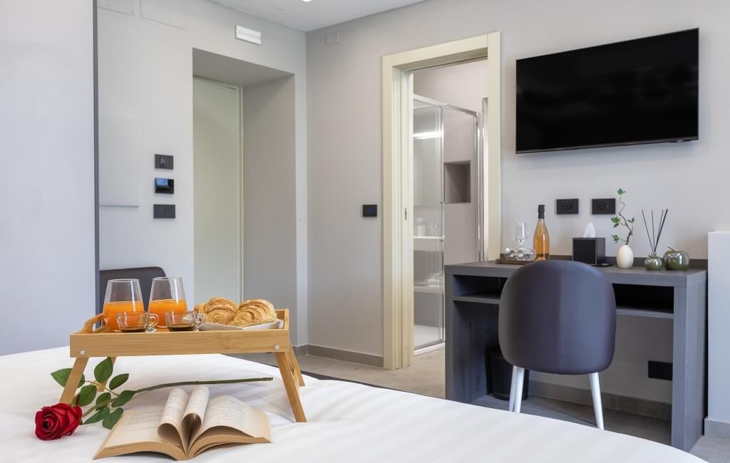 Camera d'hotel serena con letto, colazione su vassoio, scrivania grigia e TV, tonalità bianco e grigio minimalista.