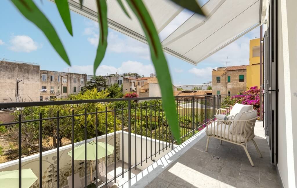 Balcone sereno con sedie bianche, tavolo, pianta verde e vista su un giardino lussureggiante e cielo blu.