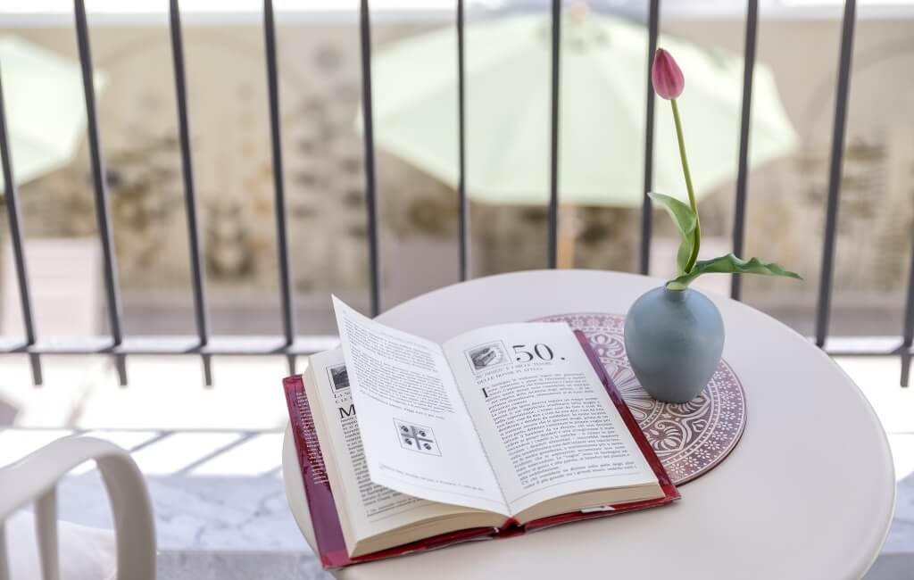 Un tavolo bianco con un libro aperto e un vaso con un tulipano rosa su un balcone con vista sfocata.