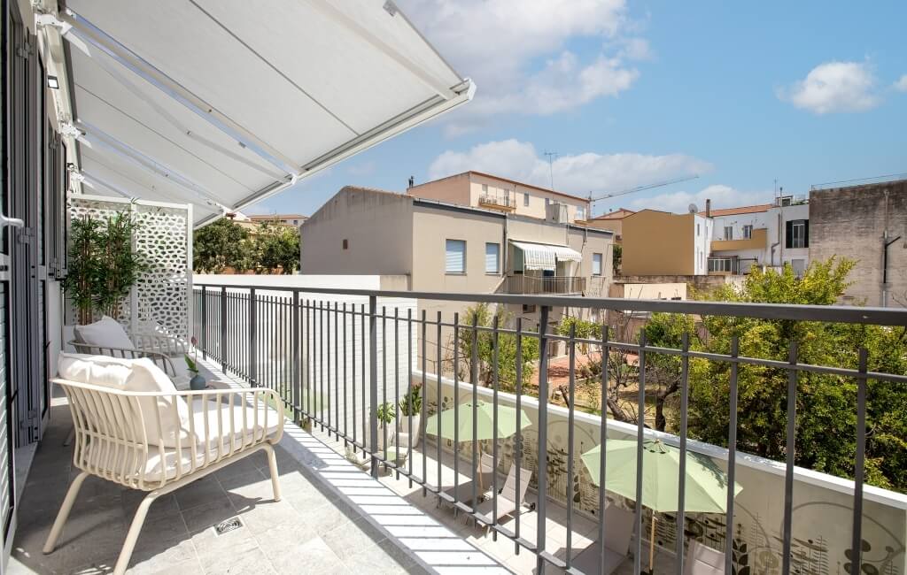 Balcone sereno con tenda bianca, divano in vimini e piante, circondato da edifici e vegetazione.