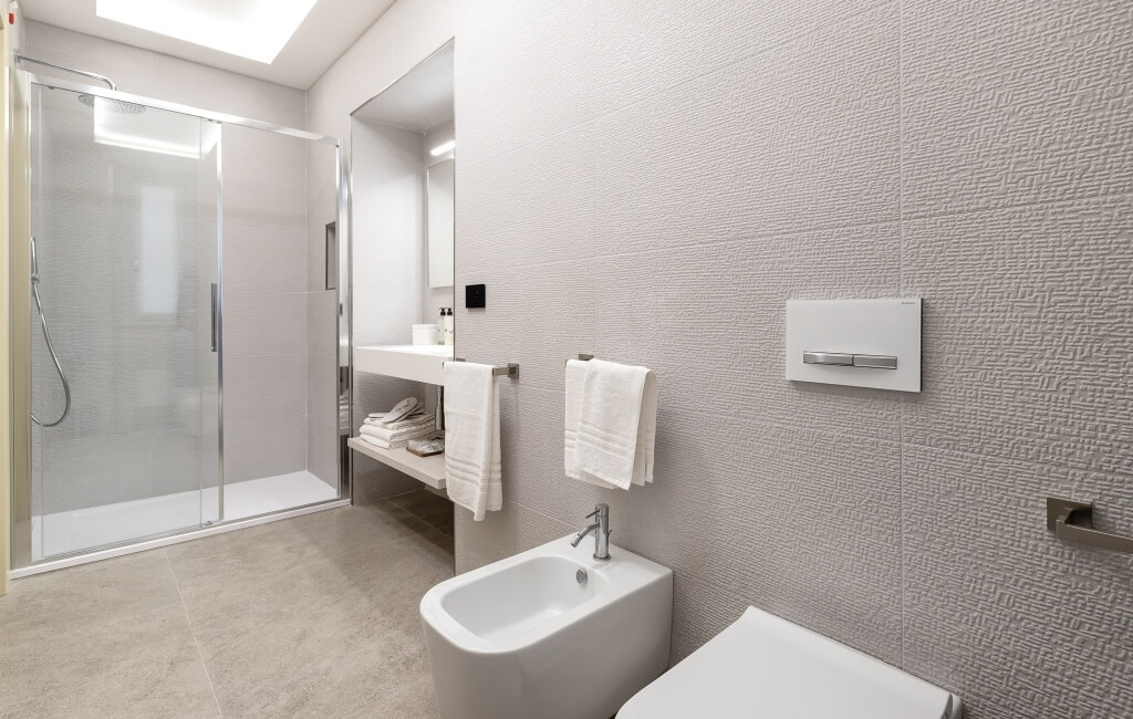 Bagno moderno bianco con doccia in vetro, lavabo, bidet e piastrelle grigie. Asciugamani piegati su mensole.