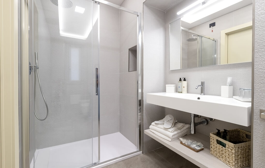 Bagno moderno con doccia in vetro a sinistra e lavabo bianco a destra, specchio sopra il lavabo e luce sopra.