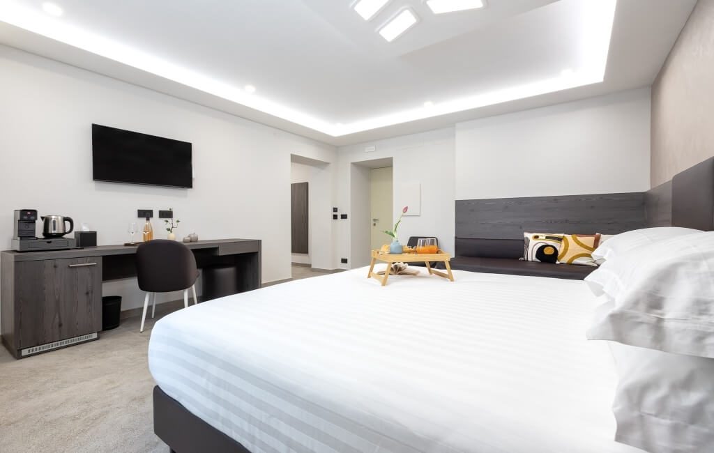 Camera d'hotel con letto, scrivania, TV, e un vassoio con cibo e bevande. Pareti bianche e pavimento chiaro.