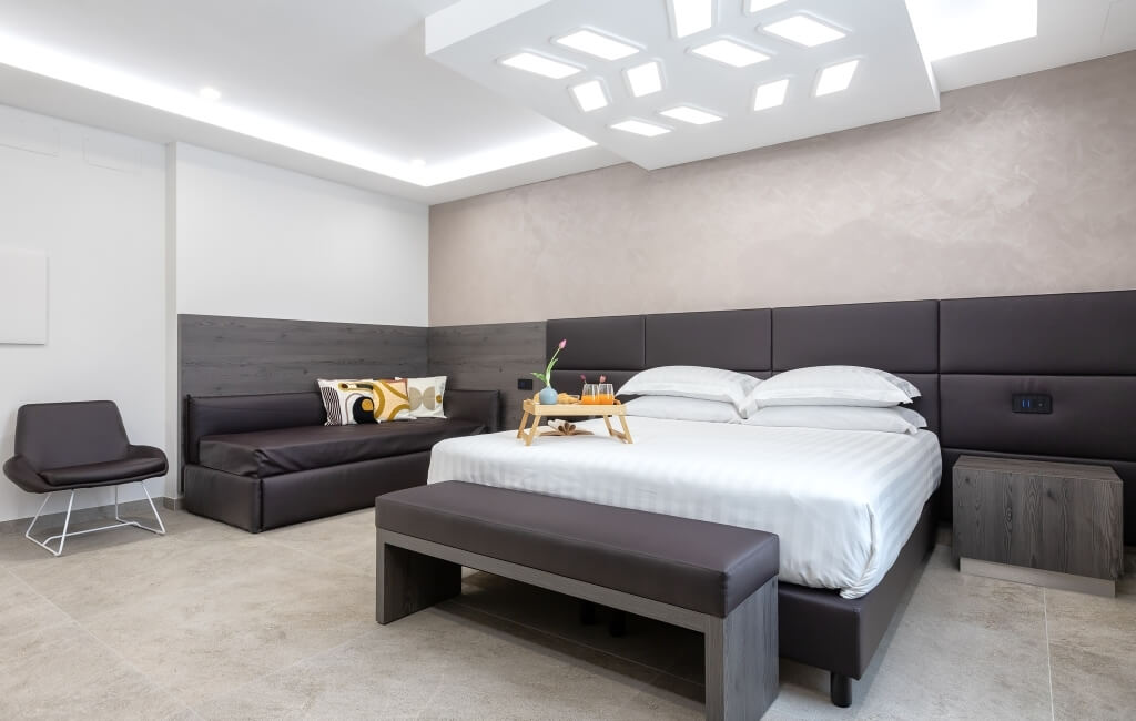 Camera d'hotel moderna con letto grande, divano, sedia e decorazioni eleganti. Muri bianchi e beige, pavimento chiaro.