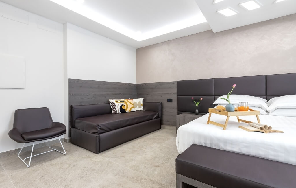 Camera d'hotel moderna con letto, divano e sedia, illuminata, con dettagli eleganti e decorazioni minimaliste.