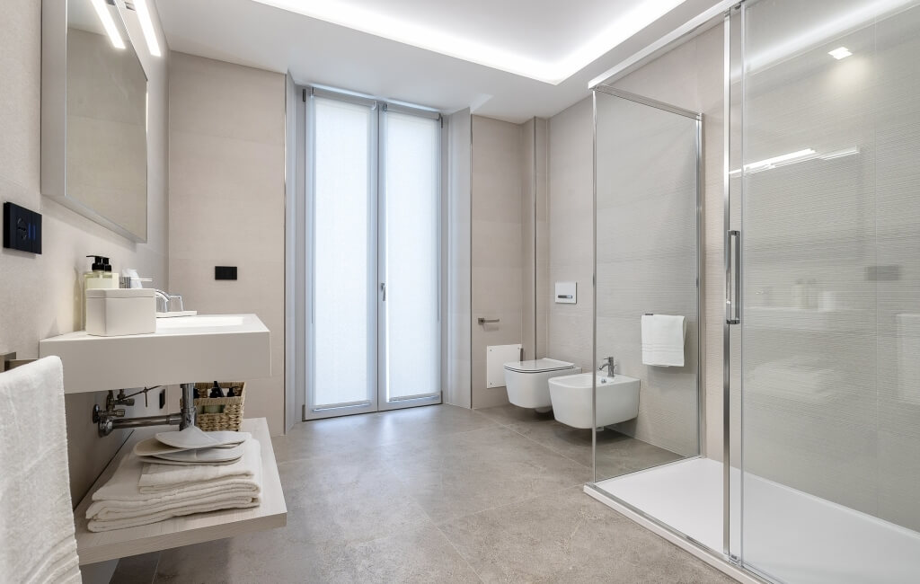 Bagno moderno e minimalista con vanità bianca, specchio grande, piastrelle grigie e finestra con vetro smerigliato.