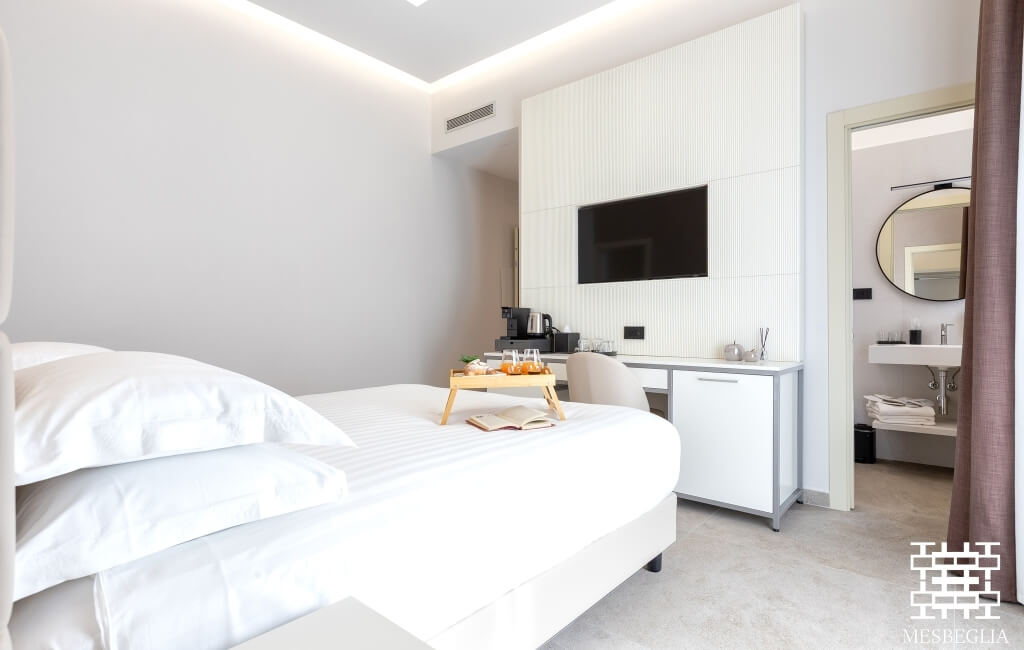 Camera d'hotel serena con tonalità bianche e beige, letto con biancheria bianca, TV, scrivania e bagno moderno visibile.