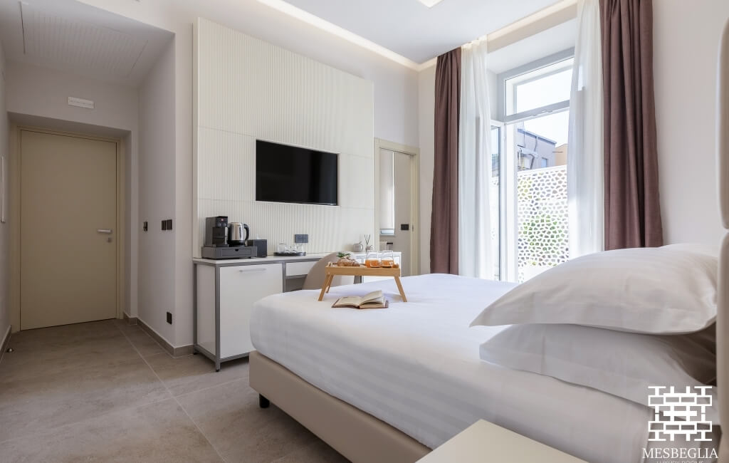 Camera d'hotel luminosa con letto, angolo cottura e TV, finestra con tende marroni e bianche.