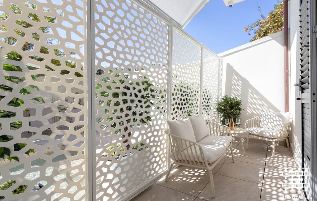 Area esterna serena con mobili bianchi in rattan e schermi a graticcio geometrici che creano separazione e ombre intricate.