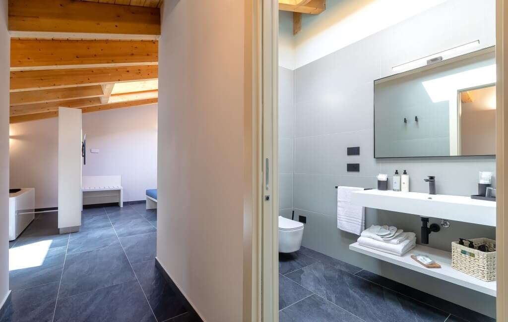 Bagno moderno con lavabo, specchio e wc, adiacente a una stanza con panca e sedia blu. Muri bianchi e pavimento grigio.