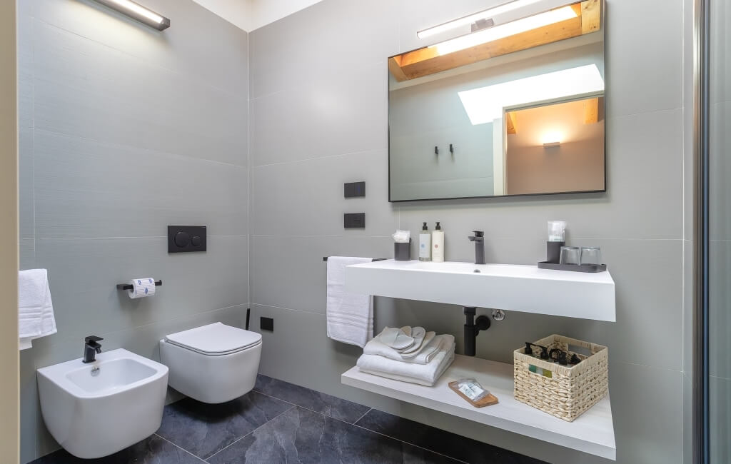 Bagno moderno minimalista con piastrelle grigie, lavabo bianco, specchio illuminato, asciugamani e cesto di vimini.