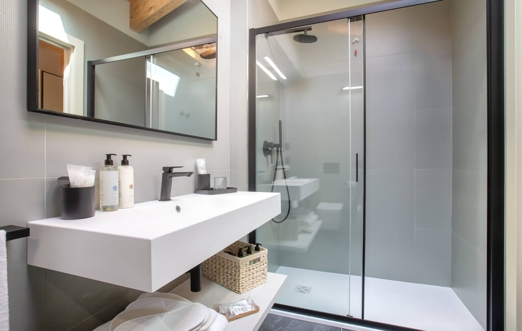 Bagno moderno con lavabo a parete, specchio rettangolare, doccia in vetro e accessori sul piano.