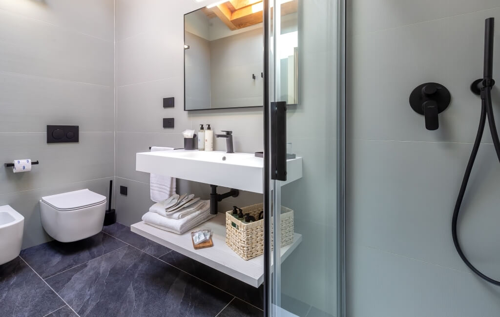 Bagno moderno minimalista con pareti grigie, pavimento scuro, lavabo e WC bianchi, doccia in vetro e accessori neri.