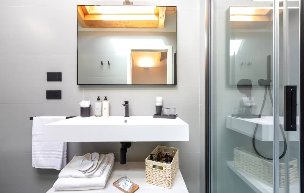 Bagno moderno minimalista con lavabo bianco, specchio rettangolare, luci sopra e doccia con porta in vetro.