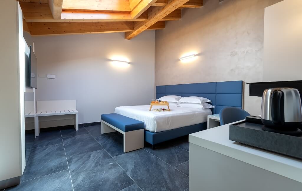 Camera d'hotel moderna con letto, panchina e scrivania, illuminata da luce naturale. Letto con testiera blu e coperta bianca.