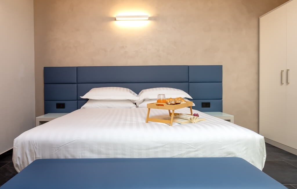 Camera d'hotel serena con letto grande, testiera blu, lenzuola bianche, vassoio con succo d'arancia e libro.