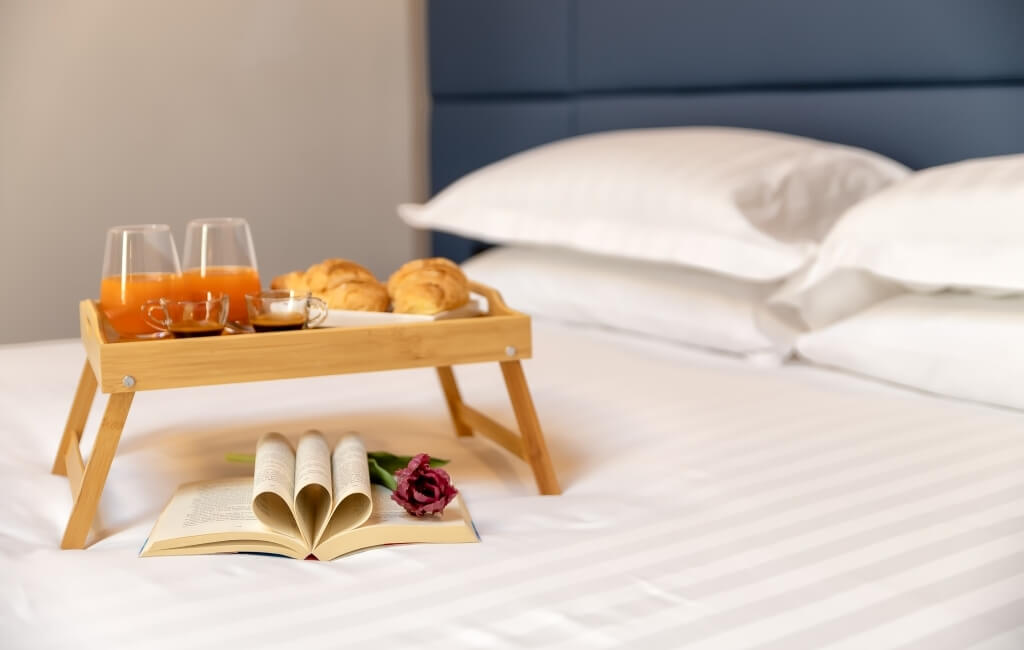 Colazione serena su un letto: vassoio con succhi, croissant e bevande, libro aperto e fiore rosa. Atmosfera rilassante.
