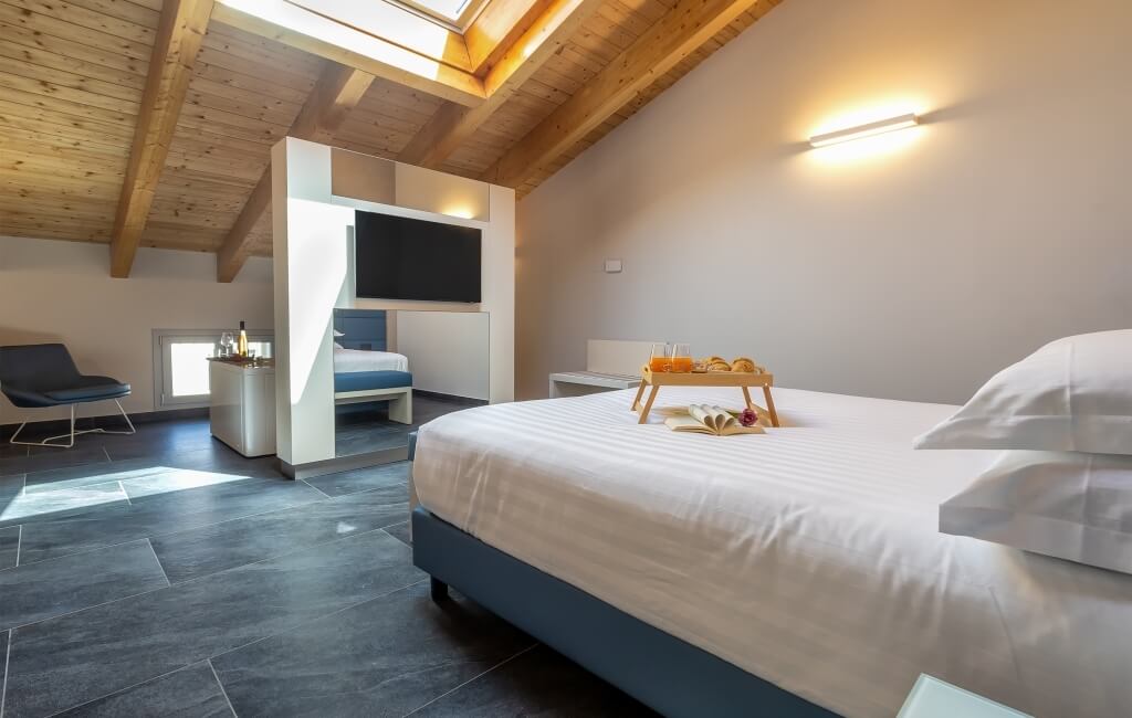 Camera d'hotel serena con letto bianco, accenti blu, TV a parete e area salotto illuminata dalla luce naturale.