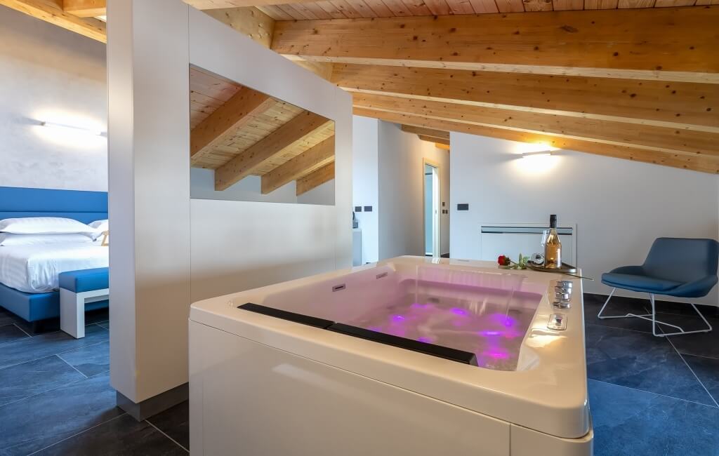 Camera d'hotel moderna con vasca bianca illuminata, vino e rose, atmosfera di lusso e relax. Letto blu sullo sfondo.