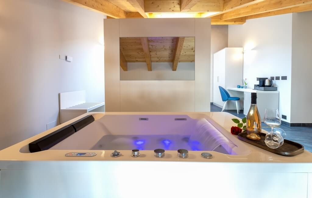 Bagno minimalista con vasca bianca, luci blu, vino, bicchieri e rosa, soffitto in legno e specchio riflettente.