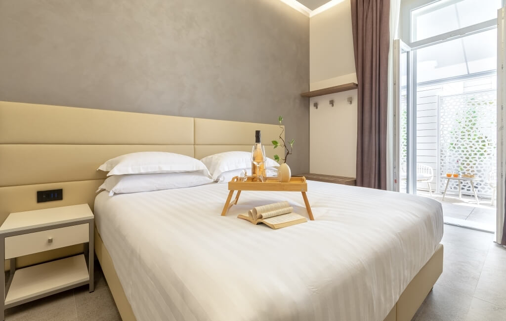 Camera serena con letto bianco, vassoio con vino e libro, comodino elegante e pareti chiare.