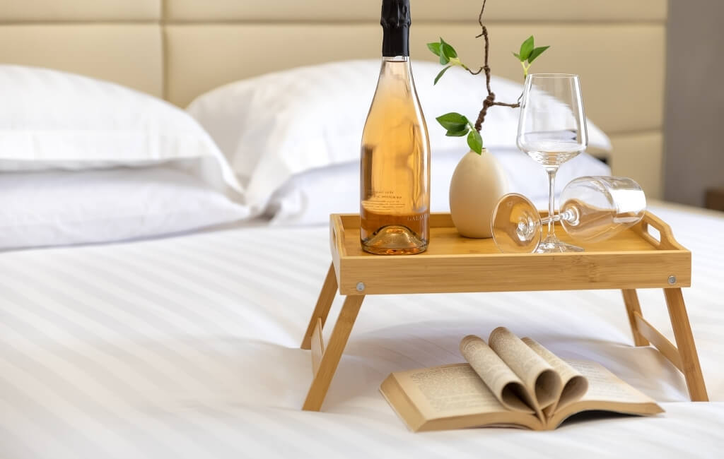 Una bottiglia di vino, due bicchieri e un libro aperto su un letto, tutto su un vassoio di legno con un vaso di fiori.
