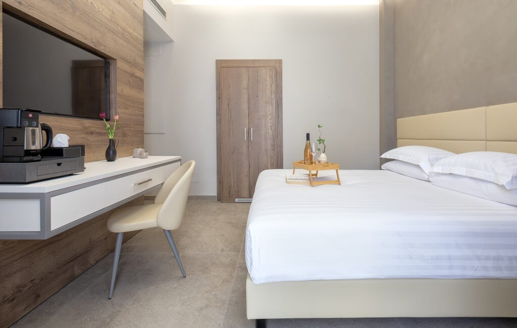 Camera d'hotel moderna con letto, scrivania e TV. Letto con coperta bianca, vassoio con vino e piante, scrivania con caffettiera.