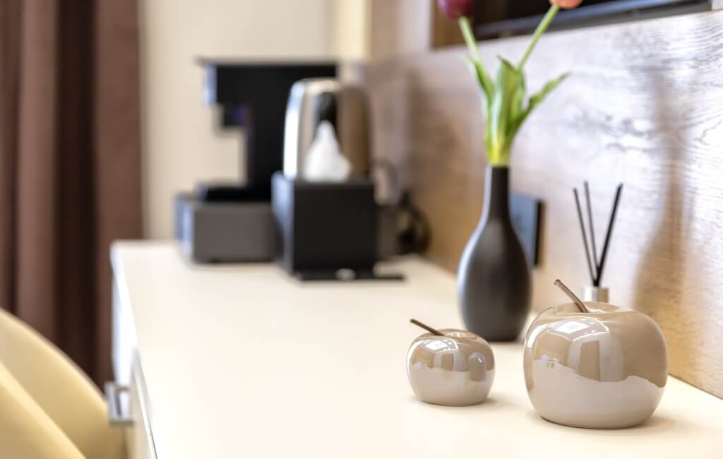 Piano di crema con diffusori a forma di mela e un vaso nero con foglie verdi e un fiore rosso, sfondo sfocato di un hotel.