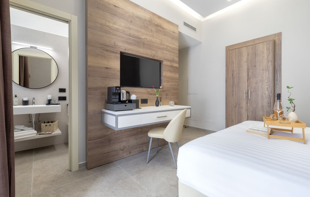 Camera d'hotel serena con tonalità bianche e legno, bagno a sinistra, scrivania, TV, letto a destra e armadio.
