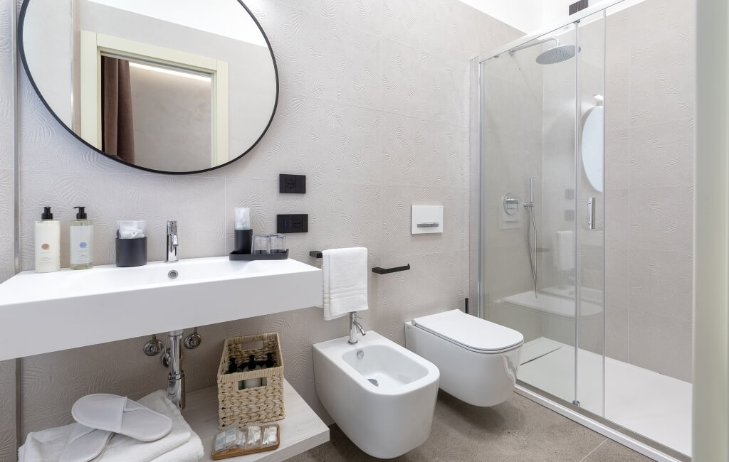 Bagno moderno con lavabo bianco, specchio rotondo, bidet, WC e doccia in vetro. Accessori in bianco e nero.